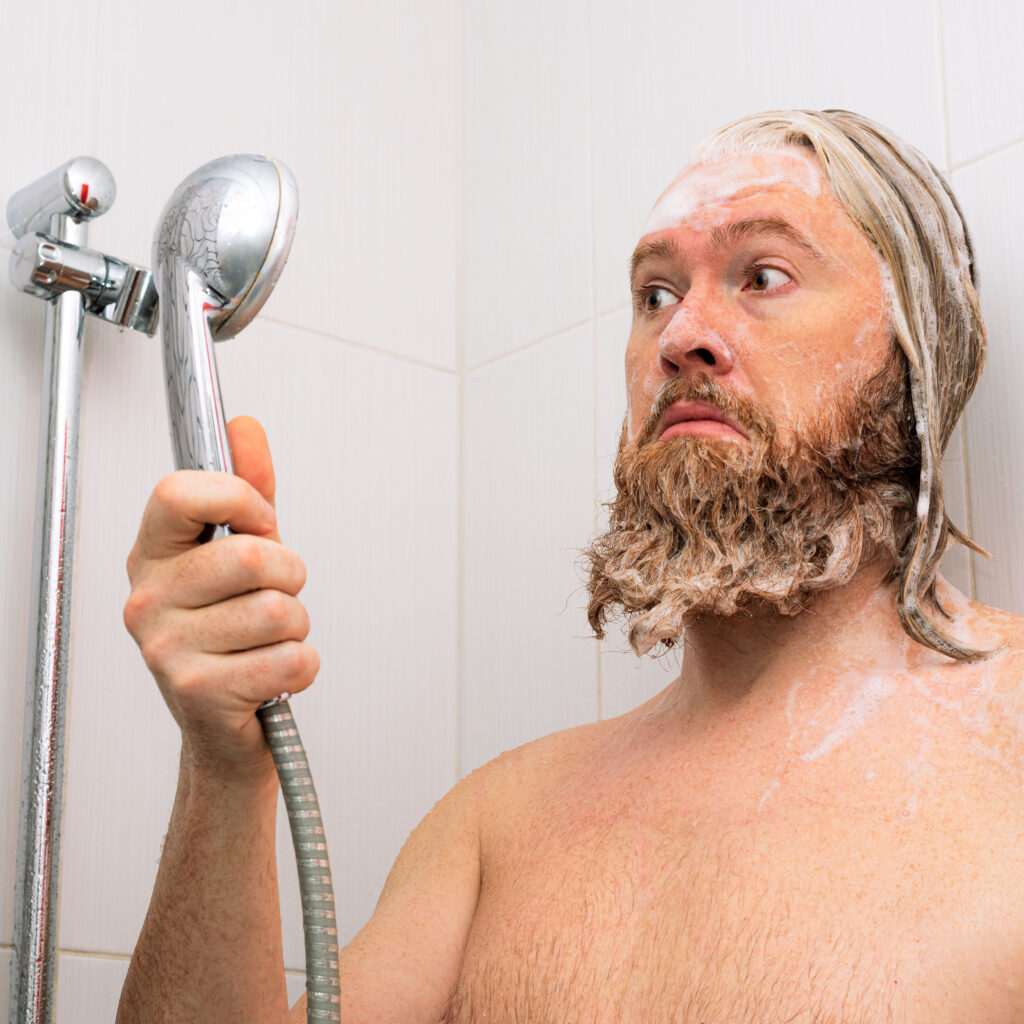 Confused bearded man with soapy head standing in the bathroom and looking at the shower while the water supply has stopped. No water coming from tap