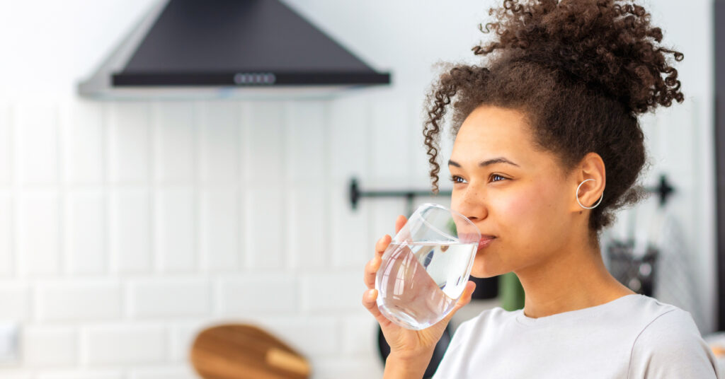 Healthy lifestyle concept. Beautiful young Afro American woman drinking clean water standing at home in the kitchen.